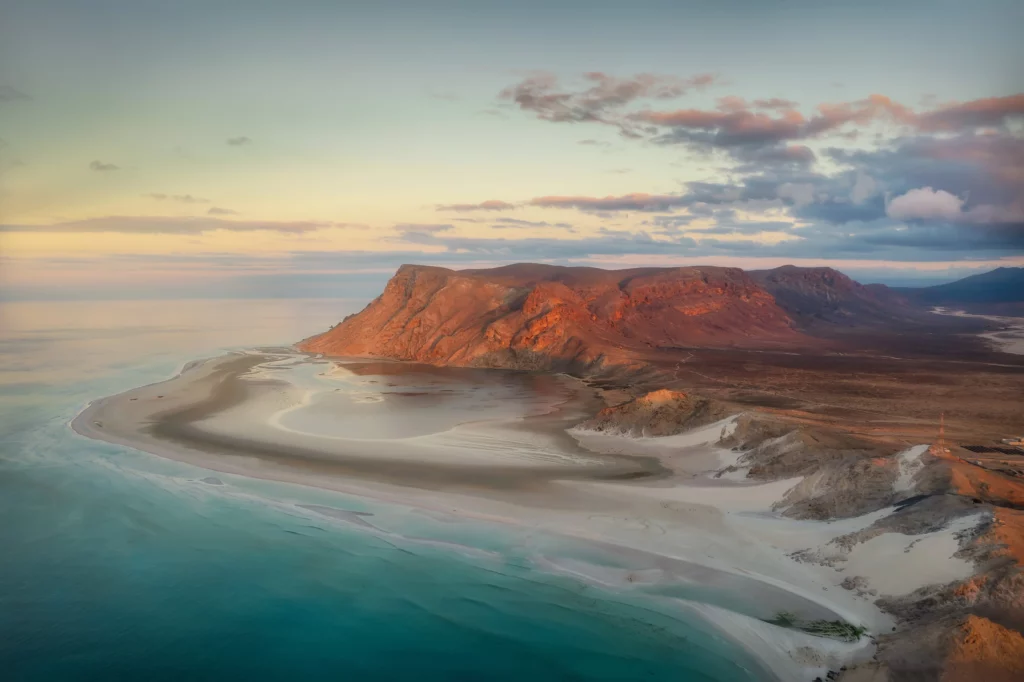 Aerial view of a sandy peninsula and dramatic red cliffs at sunset beside clear turquoise sea